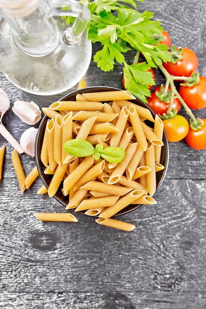 Whole grain flour penne pasta in a bowl tomatoes garlic vegetable oil in a decanter and parsley on dark wooden board background from above