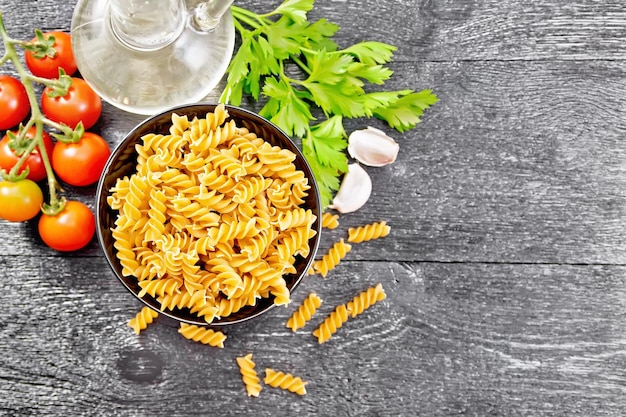 Whole grain wheat flour fusilli pasta in a bowl tomatoes garlic vegetable oil in a decanter and parsley on wooden board background from above