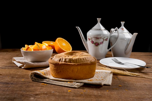 Whole orange cake on wood with orange zest, orange pieces in white jar and cake in the background.