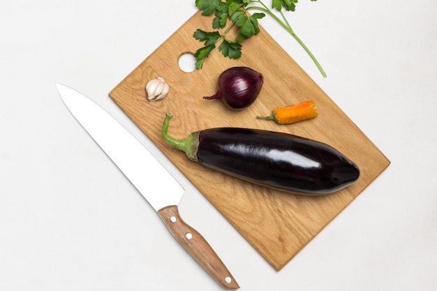 Whole raw eggplant and onions on cutting board Kitchen knife and sprig of parsley on table