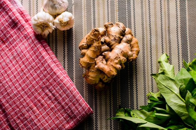 Whole raw fresh ginger place on a kitchen mat surrounded by kitchen towel whole garlic and some herbs