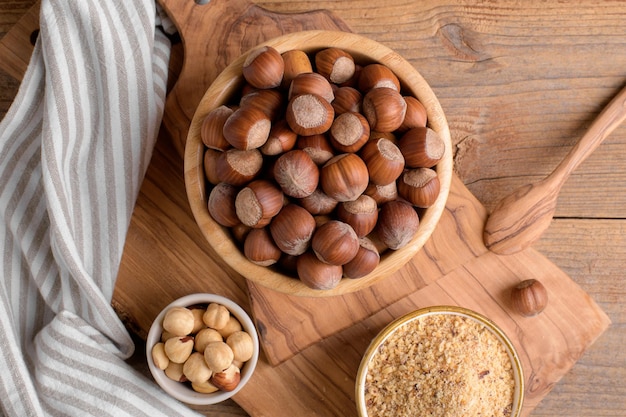 Whole raw hazelnut in a wooden bowl with peeled hazelnuts and nut flour on textured wooden background top view