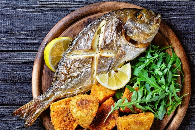 Whole roasted dorado fish with skin on baked potato coated in breadcrumbs served on a wooden cutting board with lemon wedge and fresh arugula or wild rocket salad on a wooden background, top view