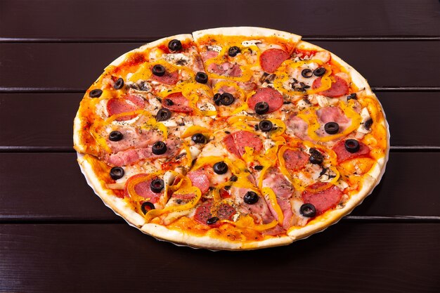 A whole rustic pizza capriciosa with sausages, bacon, pepper and mushrooms is served on a paper plate on a wooden table