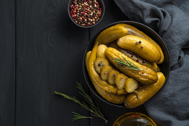 Whole and sliced pickled cucumbers with pepper and rosemary in a bowl on a wooden background. Top view, copy space.