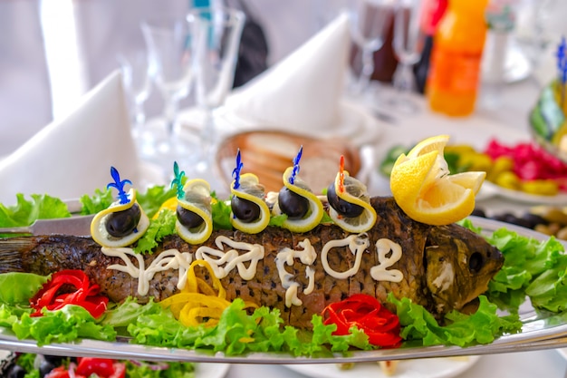 Whole stuffed fish on an oblong dish, with lemon, vegetables and olives.