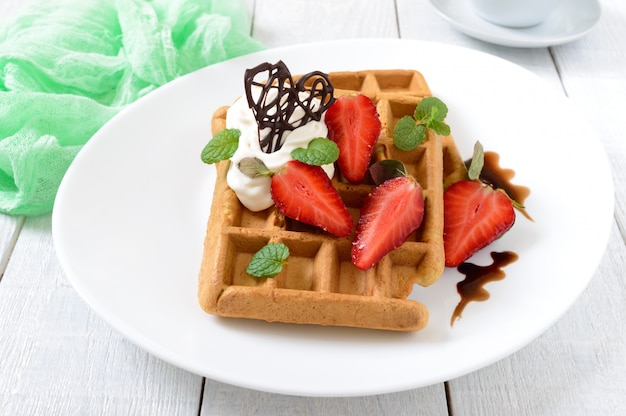 Whole wheat Belgian waffles with whipped cream, freshly chopped strawberries, mint leaves and chocolate on white wooden background. 