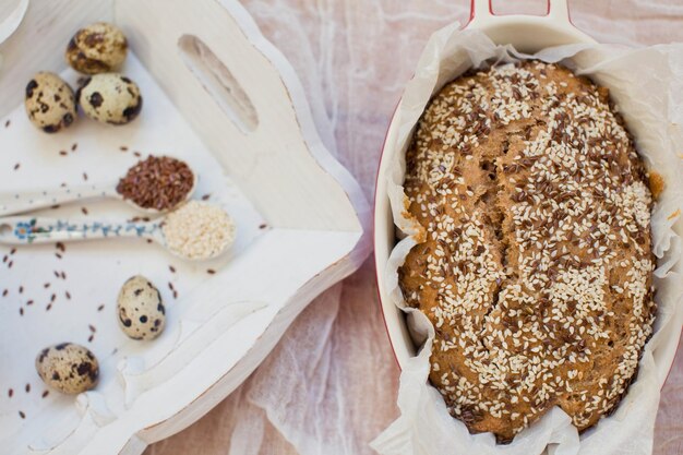 Whole wheat bread with sesame seeds and flax seeds in baking form on a light background Fresh quail eggs seeds and flour on a wooden tray