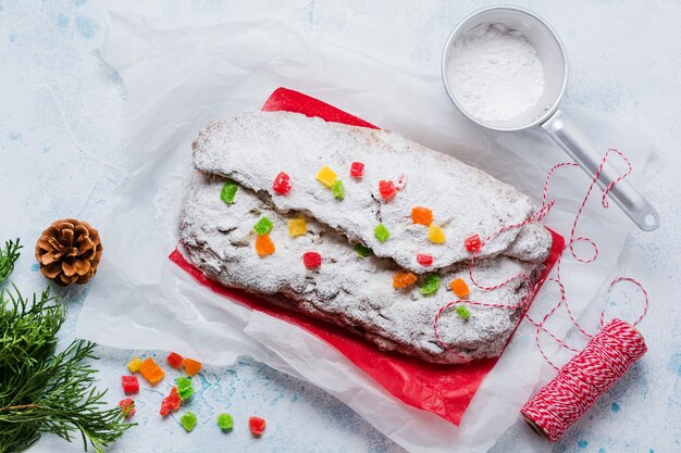 Wholegrain Stollen with raisins and powdered sugar on a linen napkin with a sieve, red ribbon over the light blue snowy concrete