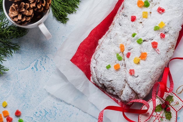 Wholegrain Stollen with raisins and powdered sugar on a linen napkin with a sieve, red ribbon over the light blue snowy concrete