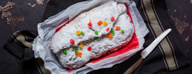 Wholegrain Stollen with raisins and powdered sugar on a linen napkin with a sieve, red ribbon over the old dark concrete background. Traditional German Christmas cake. Top view.