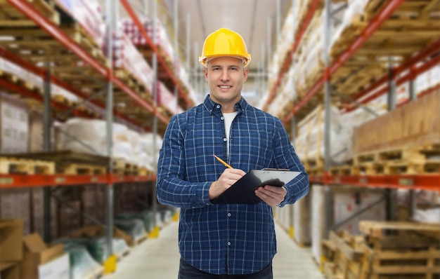 wholesale, logistic, business, export and people concept - happy man or manager in helmet with clipboard and pencil checking goods over warehouse background