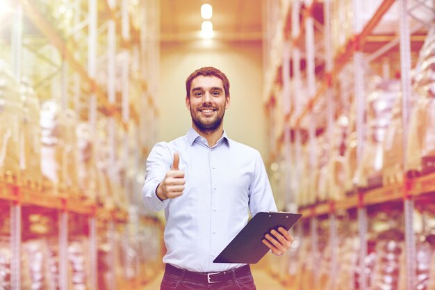 wholesale, logistic, business, export and people concept - happy man with clipboard at warehouse showing thumbs up gesture