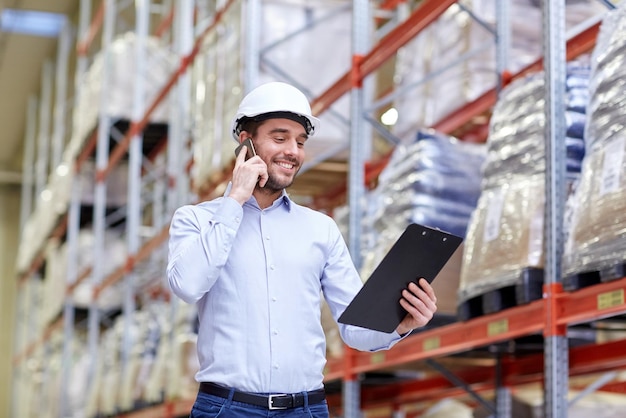 wholesale, logistic, business, export and people concept - smiling businessman with clipboard calling on smartphone at warehouse