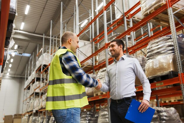 wholesale, logistic, people and export concept - manual worker and businessmen with clipboard shaking hands and making deal at warehouse