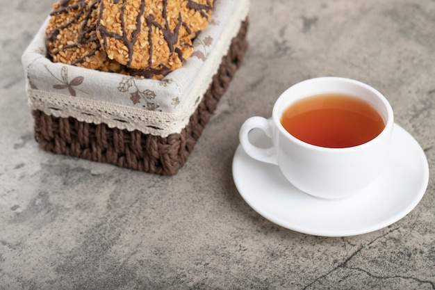 Wicker basket of oatmeal cookies with cup of black tea on marble.