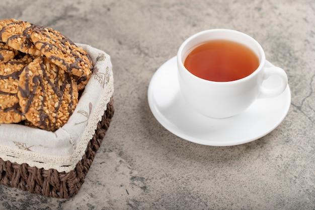 Wicker basket of oatmeal cookies with cup of black tea on marble.