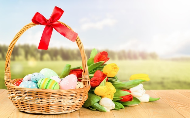 Wicker basket with painted Easter eggs and a bunch of tulips on a wooden table