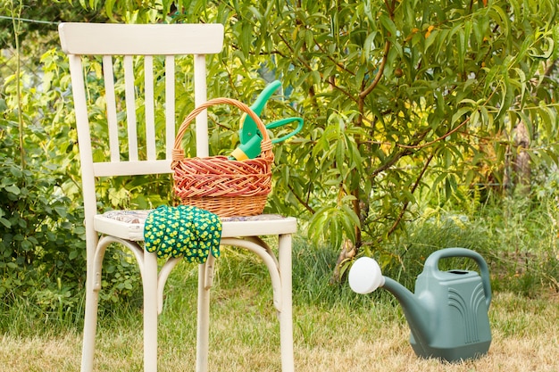 Плетеная корзина с секатором и перчатками на старом стуле, лейка на траве в естественном фоне. Садовые инструменты.