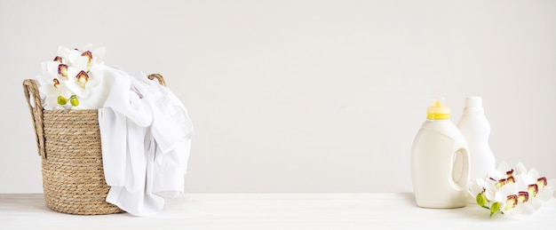 Плетеная корзина с белым бельем, гелем для стирки и смягчителем ткани на белом столе с цветами орхидеи. День стирки заголовка макета с копией пространства.