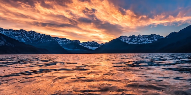 Wide angle majestic view of rocky mountains covered with snow near a lake under a cloudy sunset sky