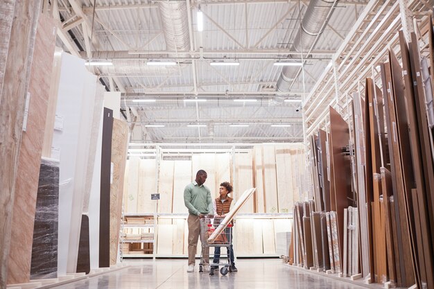 Wide angle portrait of African-American father and son shopping together in hardware store standing with cart in wood and boards isle, copy space