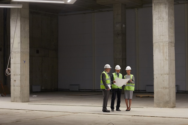 Wide angle portrait of business people wearing hardhats and holding plans while standing at construction site indoors, 