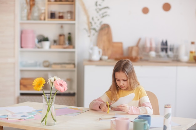 Wide angle portrait of cute girl cutting handmade holiday card for Mothers day or Valentines day while sitting at table with flowers, copy space