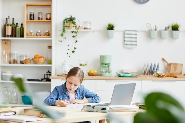 Wide angle portrait of cute smart girl writing in notebook while doing homework or studying at home in cozy interior, copy space