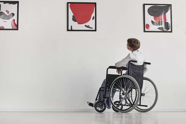 Wide angle portrait of young woman using wheelchair looking at paintings in modern art gallery, 