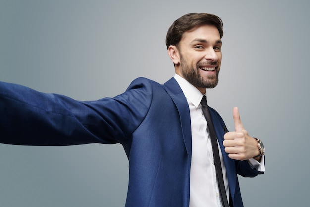 Wide angle selfie shot of young business man going thumb up on grey wall