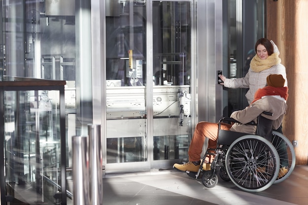 Широкоугольный вид афроамериканца в инвалидной коляске с помощью доступного лифта с молодой женщиной, копией пространства