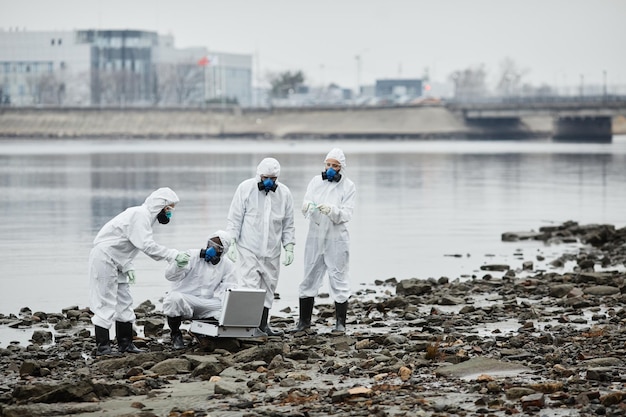 写真 化学防護服を着た人々のグループでの広角ビューは、水に有毒な廃棄物と汚染物質によってプローブを収集しています...