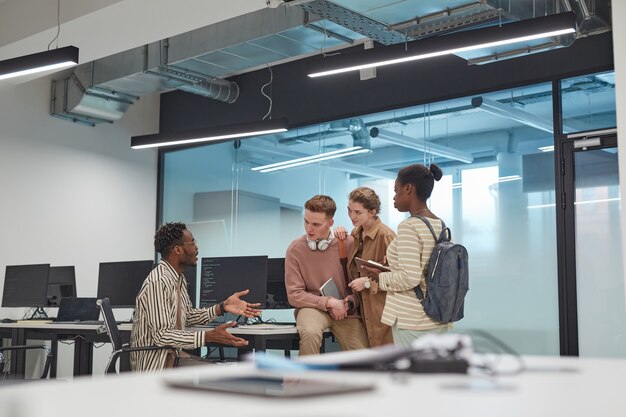 Ampio angolo di visione a diversi gruppi di giovani che discutono del progetto mentre si lavora insieme in ufficio o in un laboratorio informatico scolastico con computer, spazio di copia