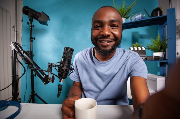 Wide angle view of famous vlogger taking a selfie hosting morning online radio show holding cup of coffe or tea. Pov shot of influencer at desk with live broadcasting setup.