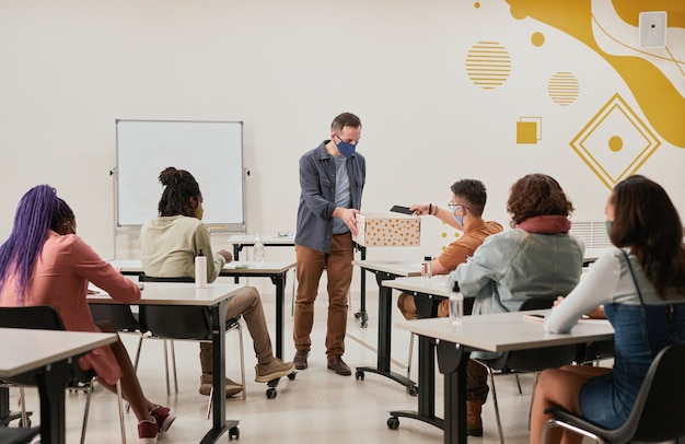 Wide angle view at male teacher collecting smartphones in school classroom with no-device policy, all wearing masks, copy space