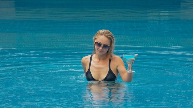 The wide middle shot pretty woman drinking blue cocktail alcohol liquor in swimming pool at hotel Portrait of sexy girl in pool outdoor