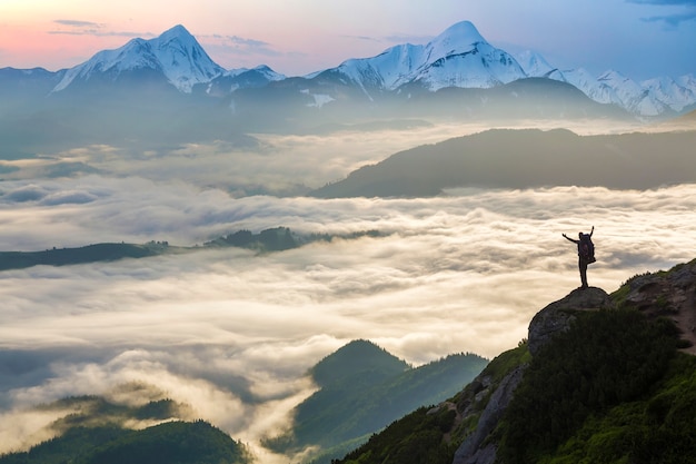 Photo wide mountain panorama. small silhouette of tourist with backpack on rocky mountain slope with raised hands over valley covered with white puffy clouds. beauty of nature, tourism and traveling concept