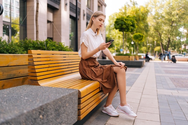 Wide shot of pretty female with long blond hair in casual clothes using smartphone sitting on bench on city street in summer sunny day