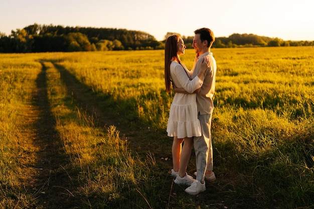 Wide shot of romantic young couple in love hugging looking at each other standing together on green meadow in summer evening during sunset with soft sunlight