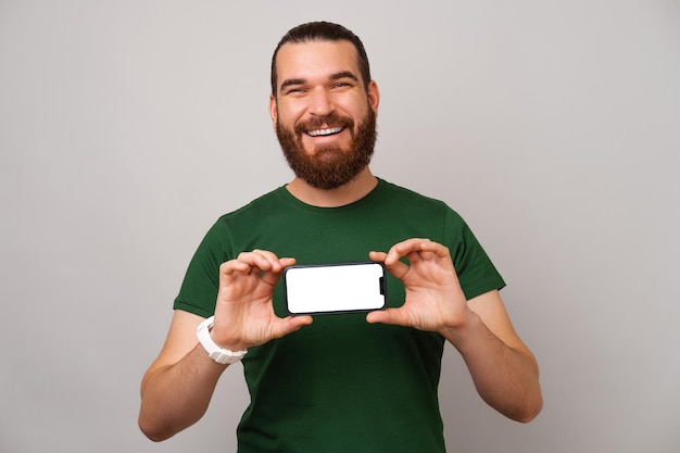 Wide smiling hipster man is holding a phone and showing its blank screen