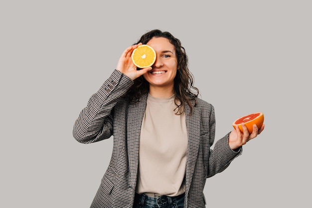 Wide smiling woman is holding a half of an orange over one eye and grapefruit