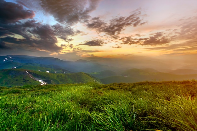 Wide summer mountain view at sunrise. Glowing orange sun raising in blue cloudy sky over green grassy hill soft grass and distant mountain range covered with morning mist. Beauty of nature concept.