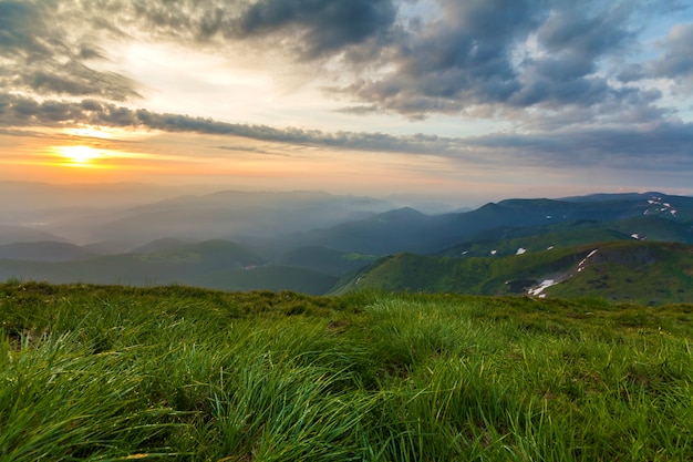 Wide summer mountain view at sunrise. Glowing orange sun raising in blue cloudy sky over green grassy hill soft grass and distant mountain range covered with morning mist. Beauty of nature concept.