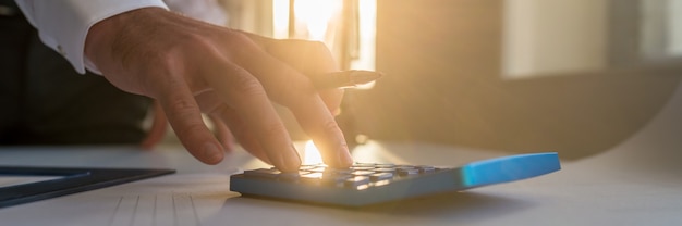 Wide view image of an architect using blue calculator holding a pencil as he works on a project with sun rays coming from the office window.