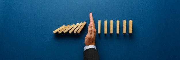 Wide view image of businessman hand stopping collapsing dominos in a conceptual image. Top view over navy blue background.