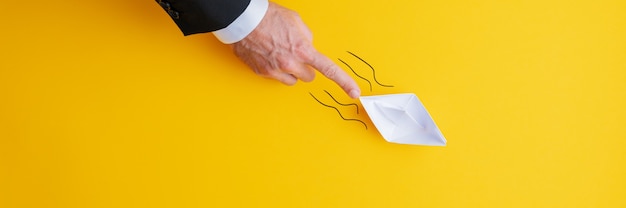 Wide view image of businessman pushing paper made origami boat forward in a conceptual image. Over yellow background with copy space.