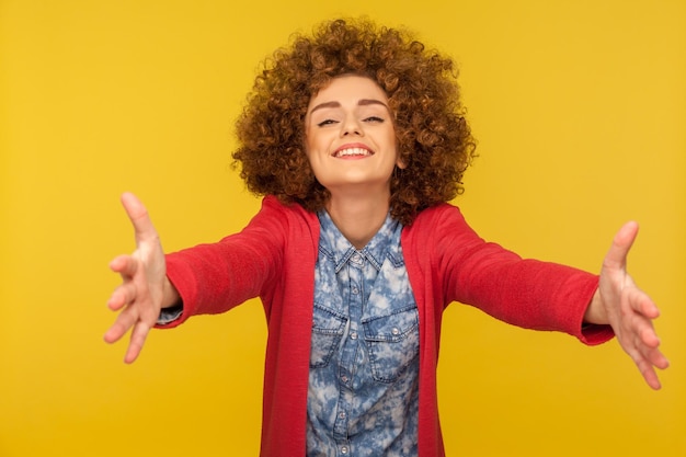 Wie wil er gratis knuffels. Portret van een vriendelijke vrouw met krullend haar die verwelkomt met wijd opgeheven handen en klaar om te omhelzen, liefde te delen, oprechte gasten te ontmoeten met een glimlach. indoor studio-opname, geïsoleerd