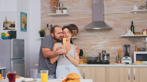 Жена поет на деревянной ложке во время танца с мужем на кухне в пижаме. Беззаботная пара смеется, веселится, веселится, наслаждается жизнью, аутентичные женатые люди, позитивные счастливые отношения