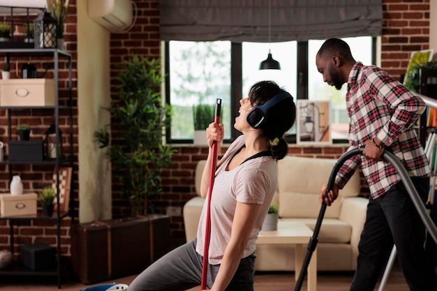 Wife sings with headphones using mop, husband is in charge of using vacuum cleaner to remove dust. Multicultural couple doing housekeeping, taking advantage of the weekend to do spring cleaning.